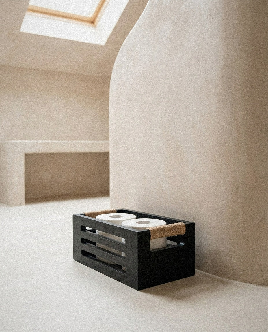 Black wood toilet paper basket with rope handles holding rolls on a plaster bathroom floor