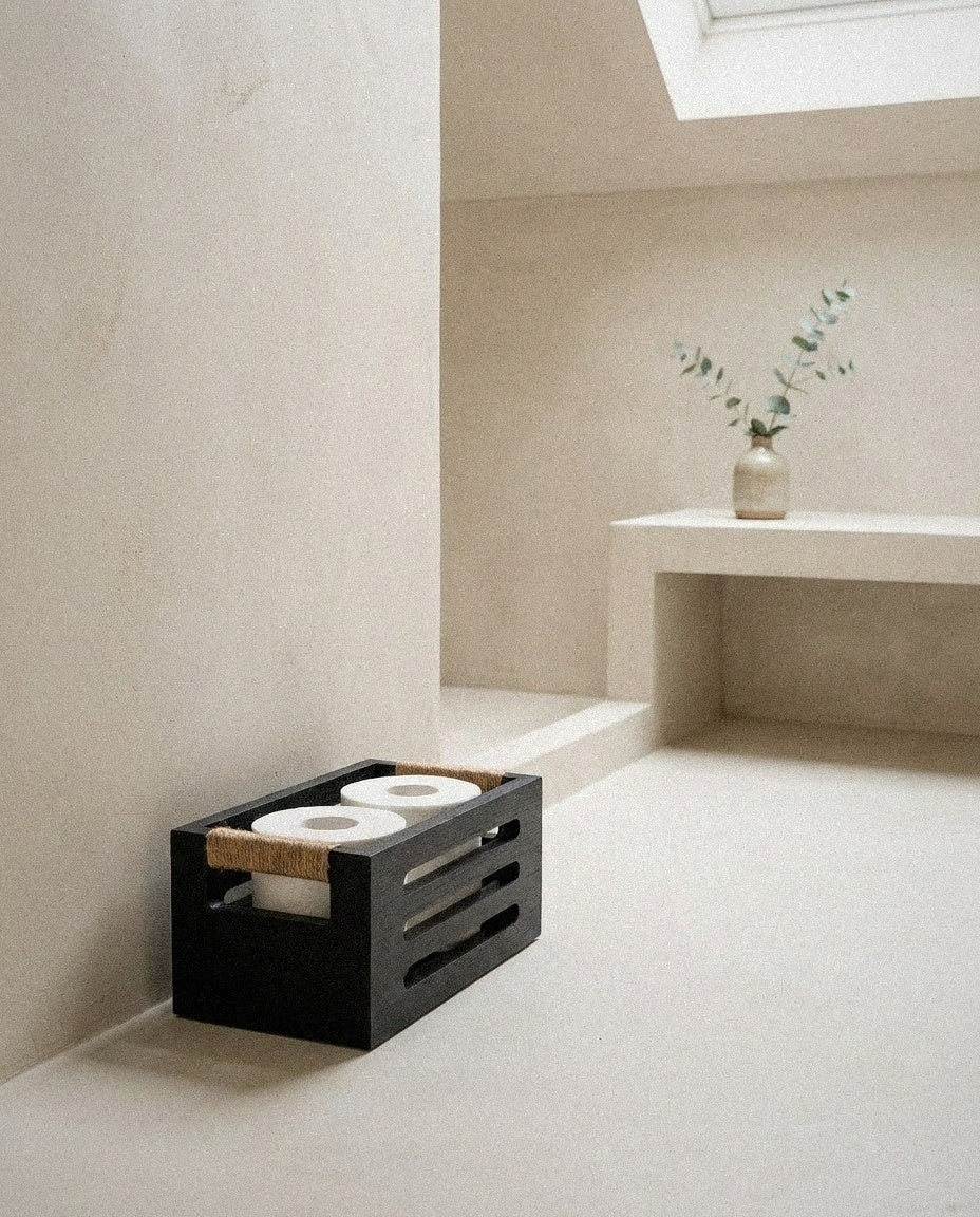 Black wooden toilet paper storage basket beside a toilet in a minimal plaster bathroom