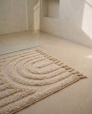 Hana cream cotton bath mat with raised rainbow arch texture and tassels on a stone floor