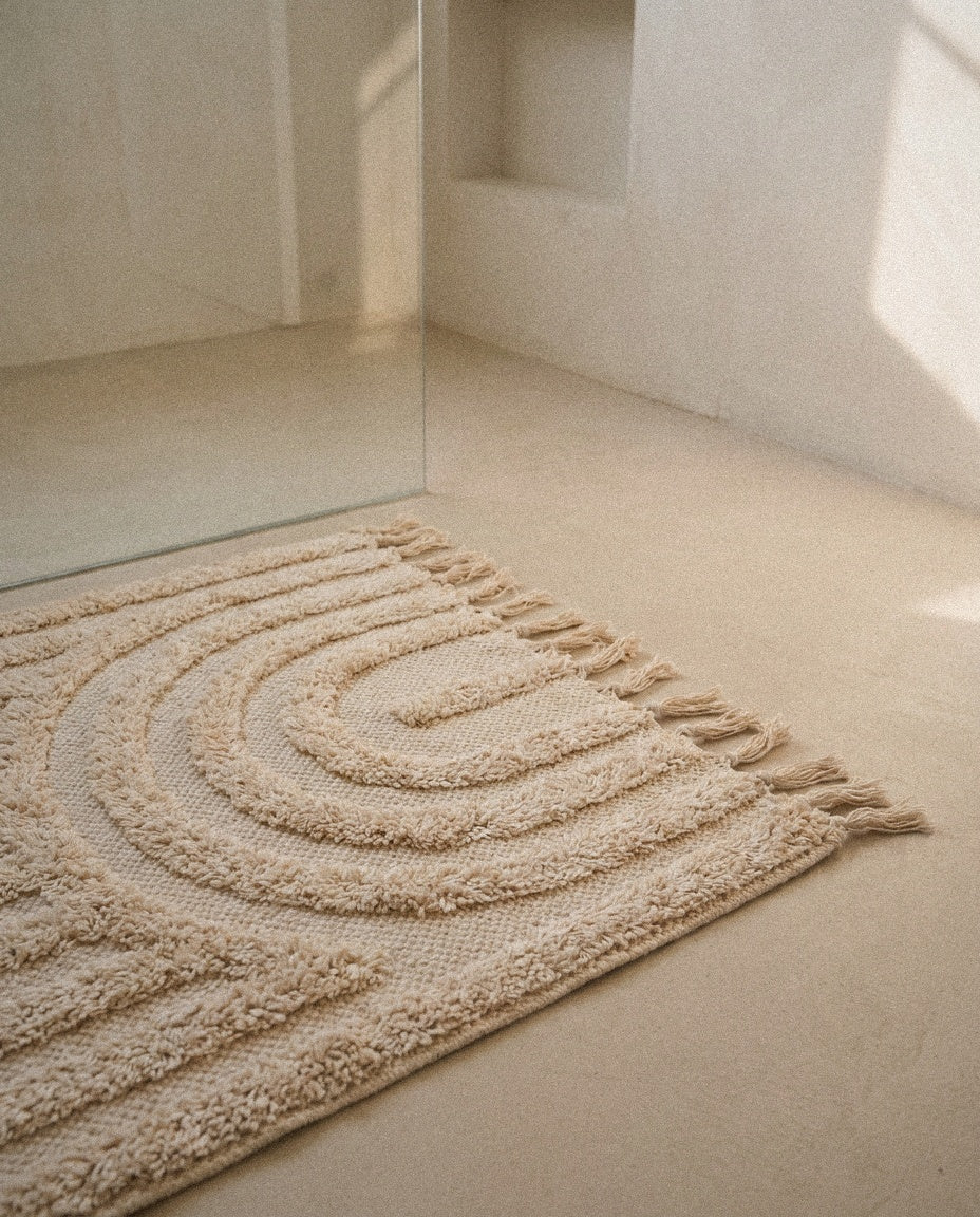 Hana cream cotton bath mat with raised rainbow arch texture and tassels on a stone floor