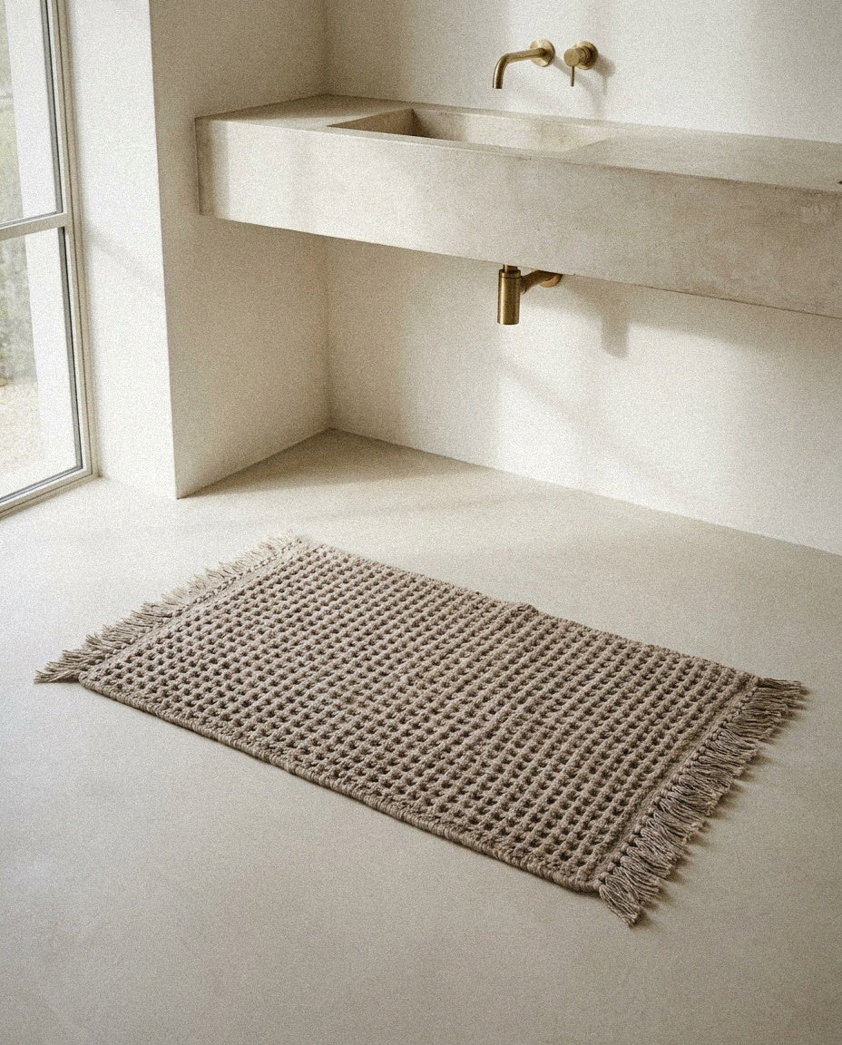Siena handwoven taupe waffle bath mat with fringe lying on a microcement bathroom floor