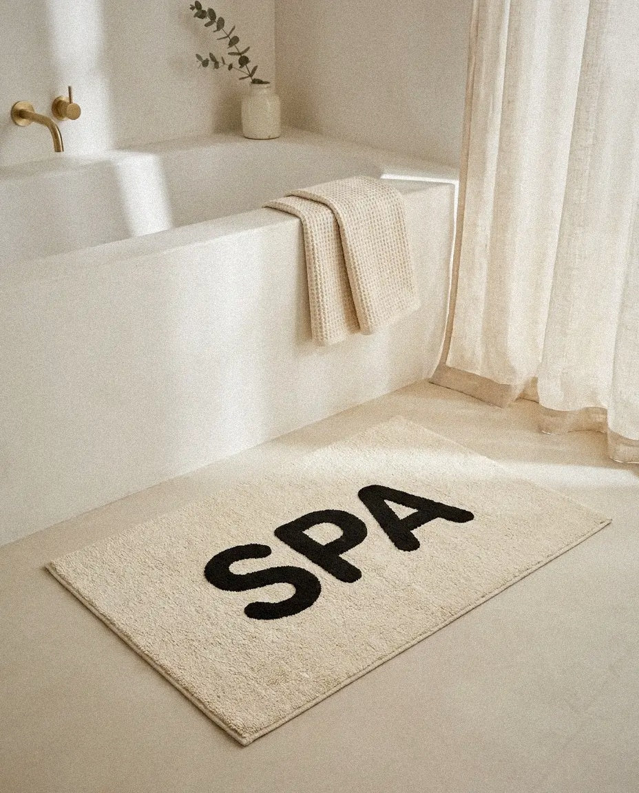Spa cotton bath mat in front of a white bathtub with a waffle towel and linen curtain