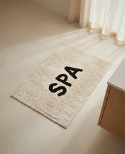 Ivory cotton SPA bath mat with black tufted lettering on a light bathroom floor in soft daylight