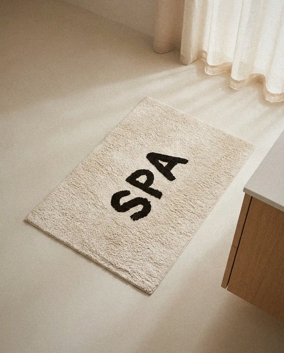 Ivory cotton SPA bath mat with black tufted lettering on a light bathroom floor in soft daylight