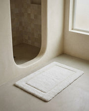 White UltraPlush cotton bath mat with raised border on a microcement floor by a curved shower