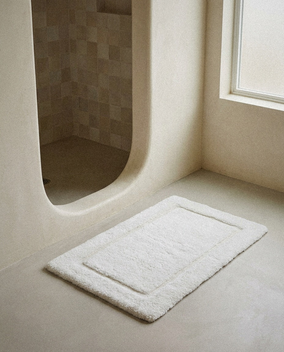White UltraPlush cotton bath mat with raised border on a microcement floor by a curved shower