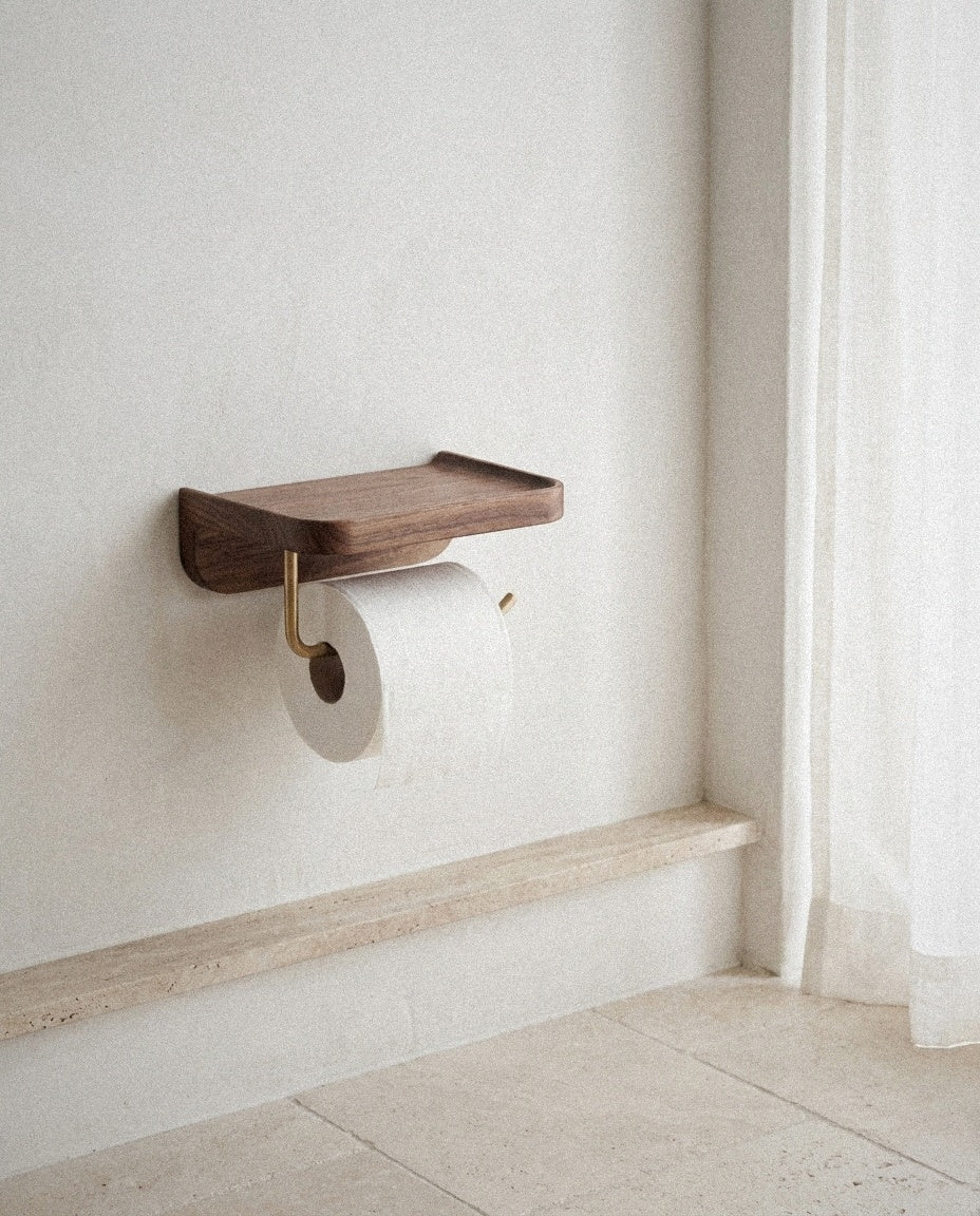 Walnut wood toilet paper holder with shelf and brass arm on a white plaster wall